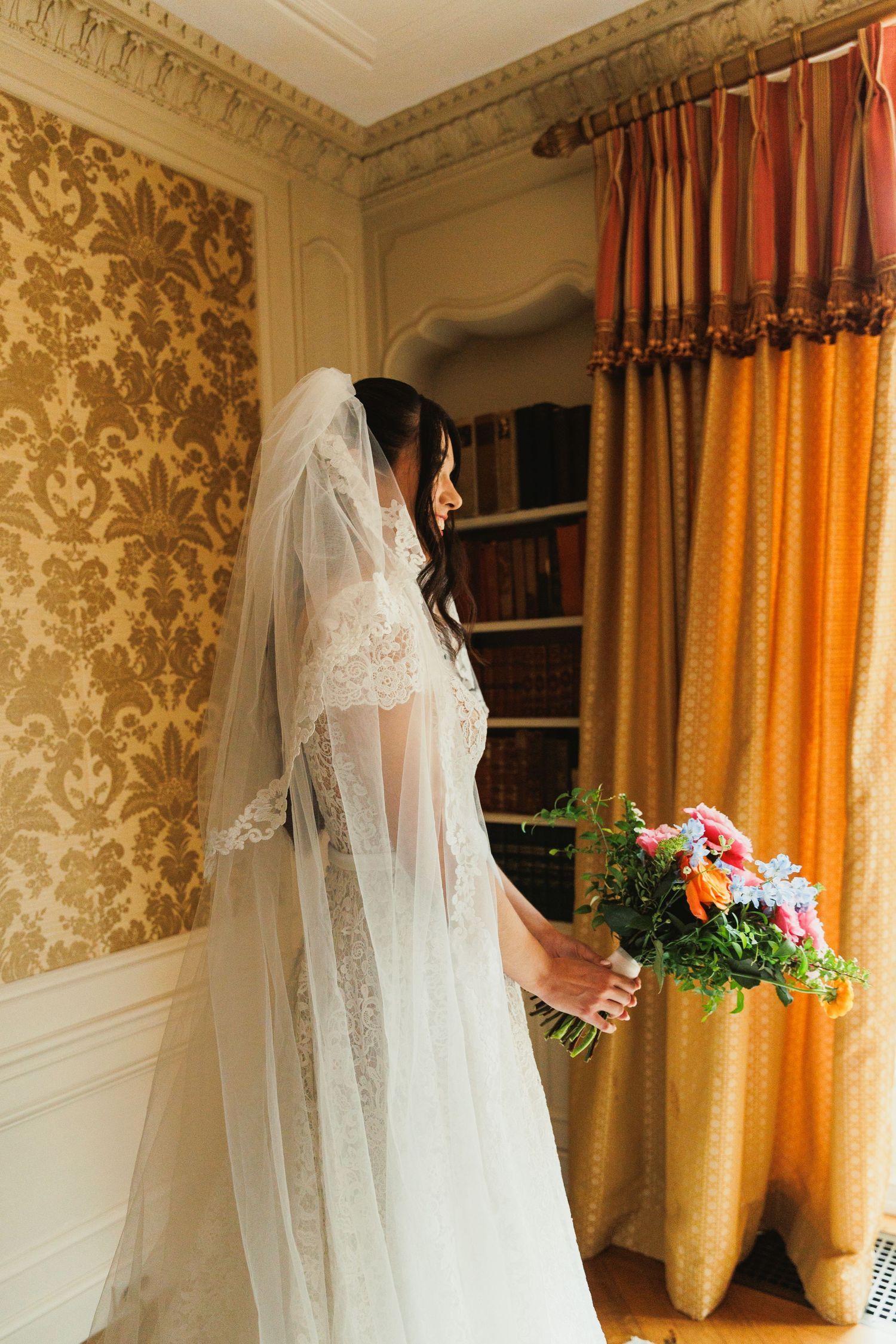 Bride in a lace veil holding her bouquet by a tall window